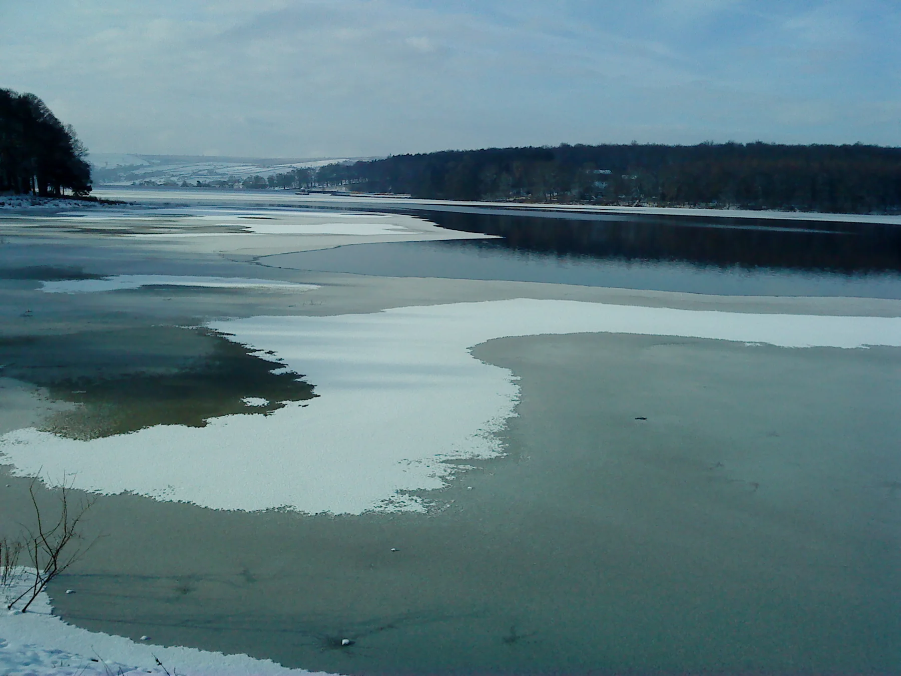 Ice and snow across Swinsty Reservoir in winter, with wooded banks and distant snowy hills