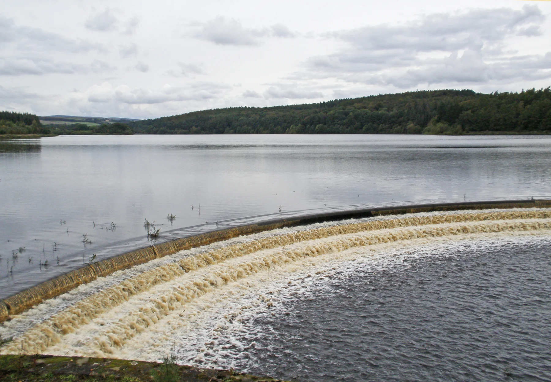 Water flowing over Fewston Reservoir’s curved spillway, with the reservoir and wooded hills beyond